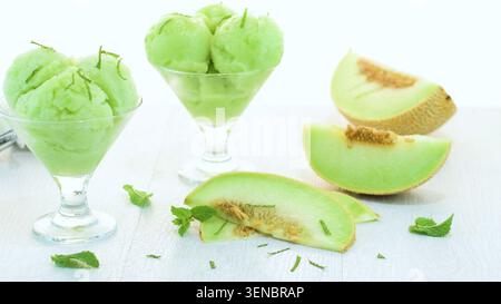 Two elegant glass dishes hold scoops of vibrant green melon sorbet garnished with mint. Fresh melon slices with seeds sit on a bright white wooden sur Stock Photo