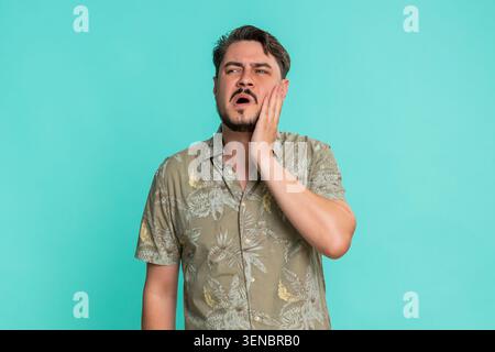 Middle-aged man holding jaw and wincing with painful expression, suffering from sharp toothache or dental issue. Young adult guy experiencing discomfort and facial tension isolated on blue background. Stock Photo