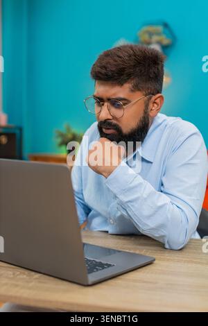 Indian man at home types on laptop stops rubs chin thinking over vital idea creative future plan. Arabian guy at table gazes screen focused eyes show deep concentration solving problem quickly today. Stock Photo
