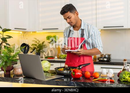 Indian man looking at laptop and writing meal notes in notebook during healthy eating study in home kitchen. Arabian guy planning diet menu and nutrition routine for weight loss learning with focus. Stock Photo
