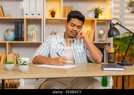 Indian man on home table writes a simple shopping list on paper and plans daily needs with calm focus. Arabian guy on home table makes notes with pen and prepares for local home store trip after work. Stock Photo