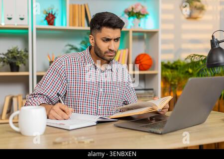 Indian man on home table reads book writes notes and checks laptop screen during focused online study in living room. Arabian guy on home table studies with calm focus in daily home learning session. Stock Photo