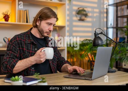 Young man on home table drinks coffee while reading laptop screen during remote work break at home office. Adult guy on home table stays focused and s Stock Photo