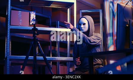Hacker wearing anonymous mask doing ransom video after gathering information from users computers using spyware software, Cybercriminal blackmailing victim in graffiti painted hideout. Stock Photo