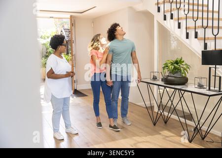 Diverse couple holding hands as woman resting on shoulder, man holding key, host gesturing in foyer Stock Photo