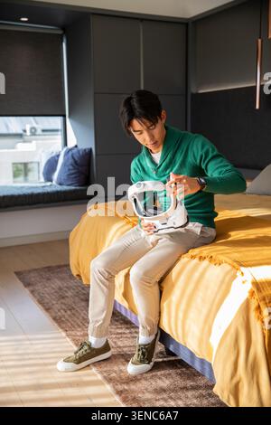 Asian man sitting on bed in modern bedroom inspecting white helmet, in green sweater and sneakers Stock Photo