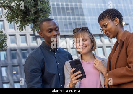 Diverse friends leaning in around phone with case, showing screen outside modern office Stock Photo