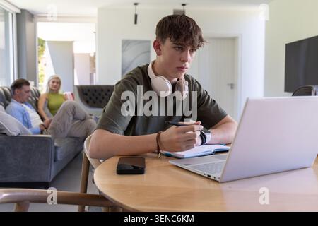 Family with teen son sitting at table using laptop headphones while parents using smartphone Stock Photo