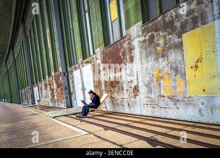Woman working on laptop in deckchair before rusted hangar gates, Tempelhof Airport, Berlin Stock Photo