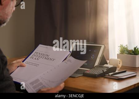 Senior man reviewing pension investment plan documents at home desk, checking financial data on tablet with calculator and paperwork. Indoor lifestyle Stock Photo