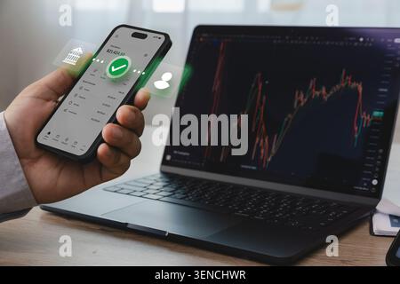 Hand holding smartphone with online banking interface beside laptop displaying financial trading chart, representing mobile money transfer, digital pa Stock Photo