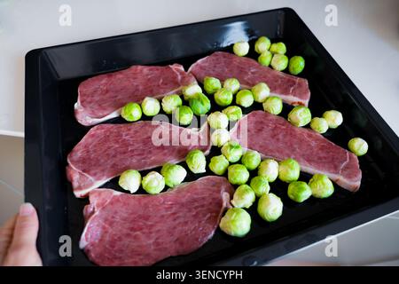 Raw pork slices and Brussels sprouts on black oven tray, healthy meal preparation Stock Photo