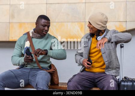 Checking watch, two men in beanie and scarf using phones and passport at station, suitcase beside Stock Photo