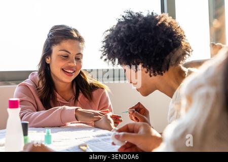 Diverse female friends sitting at table near window, one painting nails using brush and polish. Manicure, nailart, lacquer, beauty, selfcare, sunlit, Stock Photo