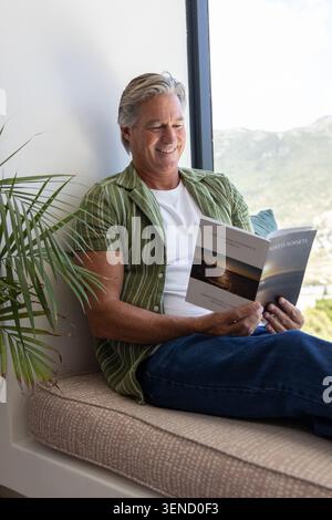 Senior man sitting on window seat reading paperback showing sunset by window with teal pillow, palm Stock Photo