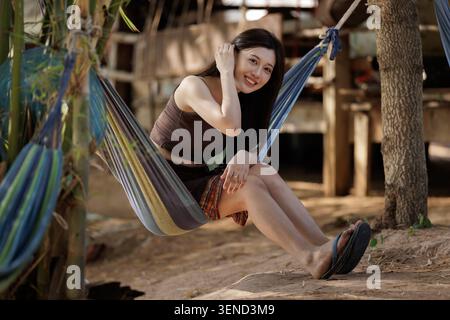 Portrait of a cheerful young woman sitting comfortably in a vibrant hanging fabric hammock in a relaxed outdoor setting. Stock Photo