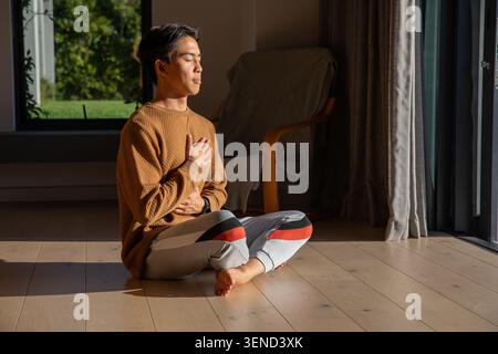Asian man sitting cross-legged doing breathwork on wood floor at home, wearing brown sweater Stock Photo