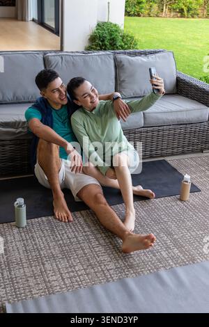 Asian couple in sportswear sitting on patio yoga mats taking selfie with phone and water bottles Stock Photo