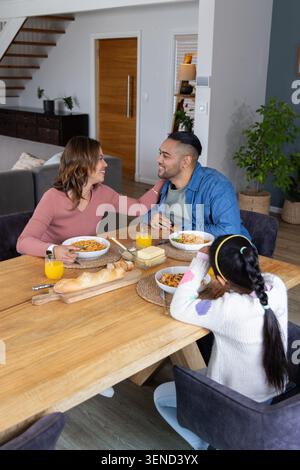 Family of three eating spaghetti in dining room at wooden table with bread, woman reaching across Stock Photo