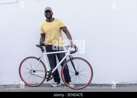 Adult African man leaning on road bicycle holding smartphone on sidewalk against white wall Stock Photo