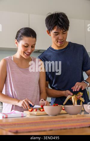 Diverse couple slicing strawberries and peeling banana at counter with beige bowls on wooden board Stock Photo