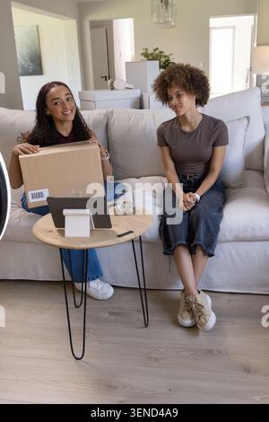 Two women sitting on sofa in bright living room, holding box and smiling beside tablet stand Stock Photo