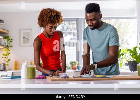African American couple arranging colored pins, sketching on wood board at kitchen island Stock Photo