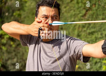 Archer concentrating, drawing bowstring with arrow, practicing sport in a green outdoor setting Stock Photo