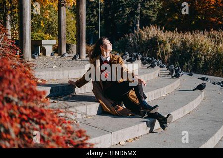 Woman sits on stone park steps surrounded by birds in sunlight. Mindful walking outdoor therapy, slow living autumn routine, connecting with nature Stock Photo
