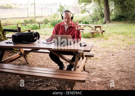 Man doing a video call from a park, working on his laptop while wearing headphones Stock Photo
