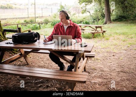 Man wearing headphones and using a laptop while writing on a notebook at a picnic table in a park Stock Photo