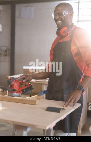 Red electric sander sanding wooden plank clamped to workbench in sunlit carpentry workshop Stock Photo