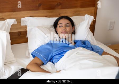 Adult woman lying in bed propped on white pillows in satin pajamas with smartphone and duvet Stock Photo