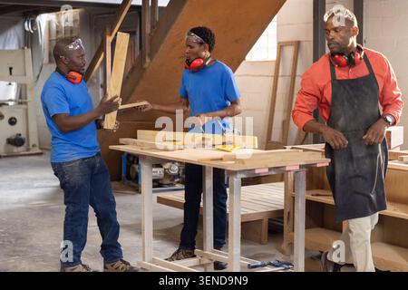 African American instructor guiding 2 apprentices passing planks in workshop wearing safety goggles Stock Photo