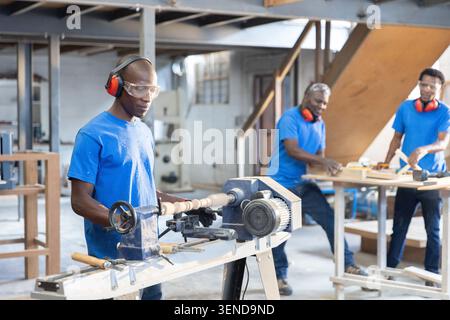 African American coworkers shaping spindle on lathe, sanding wood at workshop using safety gear Stock Photo