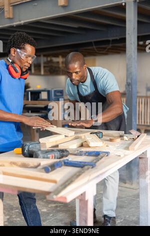 African American coworkers cutting wooden planks at workbench in woodshop using cordless drill Stock Photo
