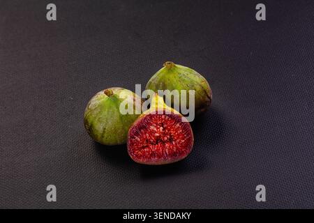 Three green figs arranged on black textured surface, one cut open to show vibrant red flesh seeds. Close up food photography with high contrast lighti Stock Photo
