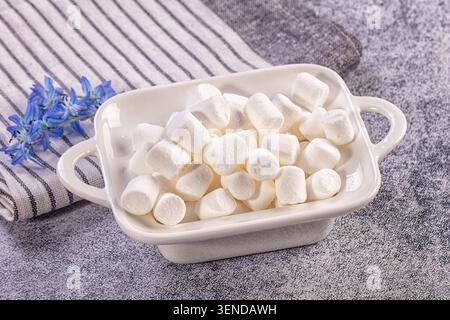 Closeup of fluffy white marshmallows piled in a white ceramic baking dish with handles, placed on a gray textured surface next to a striped cloth blue Stock Photo