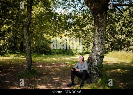 Man wearing headphones using a smartphone while sitting under a tree in nature Stock Photo