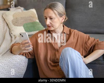 Stylish middle eged smiling woman browsing smartphone and engaging with AI-powered assistant at home. Concept of smart living, digital support, and se Stock Photo