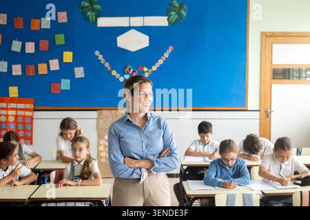 Professional elementary school teacher standing in a bright classroom while students focus on their schoolwork. Stock Photo