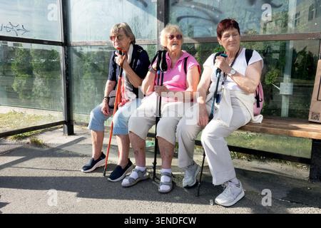 Three senior women with nordic walking poles sitting on a wooden bench inside a bus stop Stock Photo
