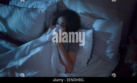Scared stressed African American woman hiding under blanket terror horror feeling stress in bed lying at night suffering with nightmare sleep disorder Stock Photo