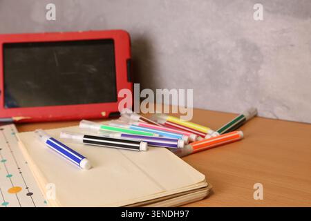 A desk shows a collection of markers in different colors scattered around a closed notebook. A tablet sits in a red case nearby, creating a casual workspace environment. Stock Photo