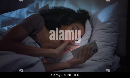 Calm peaceful African American girl woman lying in bed finishing scrolling mobile phone network at night bedroom female using smartphone nap sleep Stock Photo