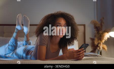 Positive African American woman chatting with mobile phone lying on bed at night girl using application smiling looking at camera relaxing at bedroom Stock Photo