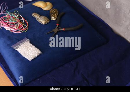 A workspace shows various crafting materials and tools on blue fabric. There are beads, a ring, pliers, and decorative items ready for use. Stock Photo