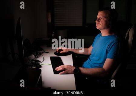 Man concentrating while using a computer at a desk in a dark room Stock Photo