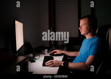 Man wearing headphones working on computer, typing on keyboard and using mouse Stock Photo