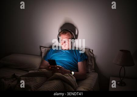 Man staying awake in bed, using a smartphone and headphones at night Stock Photo